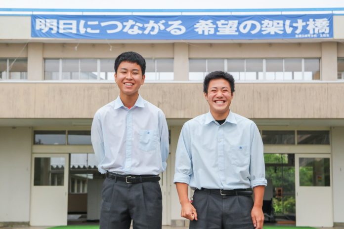 「明日につながる 希望の架け橋」と書かれた青い横断幕が掲げられた建物の前に、志津川高校の及川拓海さんと木下巧大さんが笑顔で並んで立っている写真