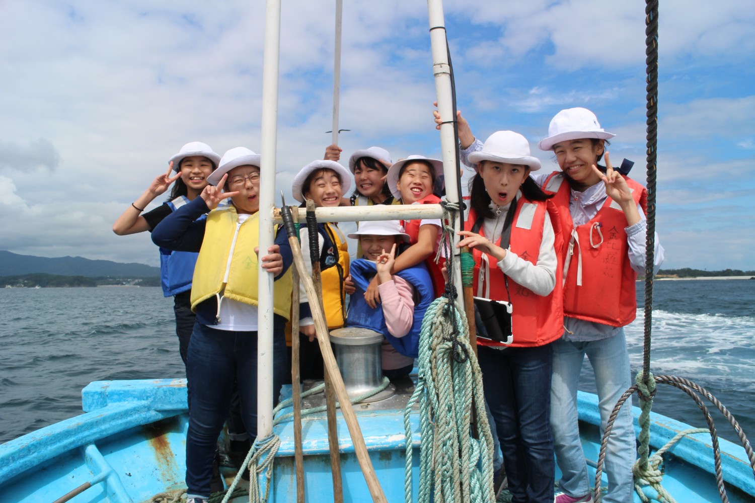青い漁船の先端部分で白い帽子とカラフルな救命胴衣を着た小学生たちが、海の風を受けながら笑顔でピースサインをしたり、楽しそうにポーズをとっている写真