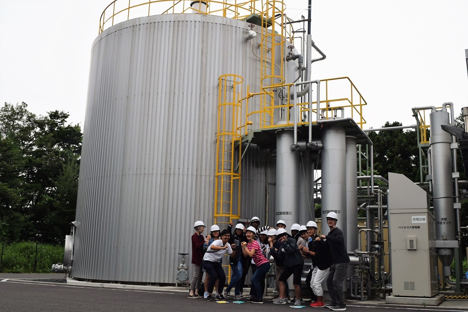 銀色の大型タンクとパイプが立ち並ぶ工場の一角で、白いヘルメットをかぶった若者たちがポーズをとって並んでいる写真