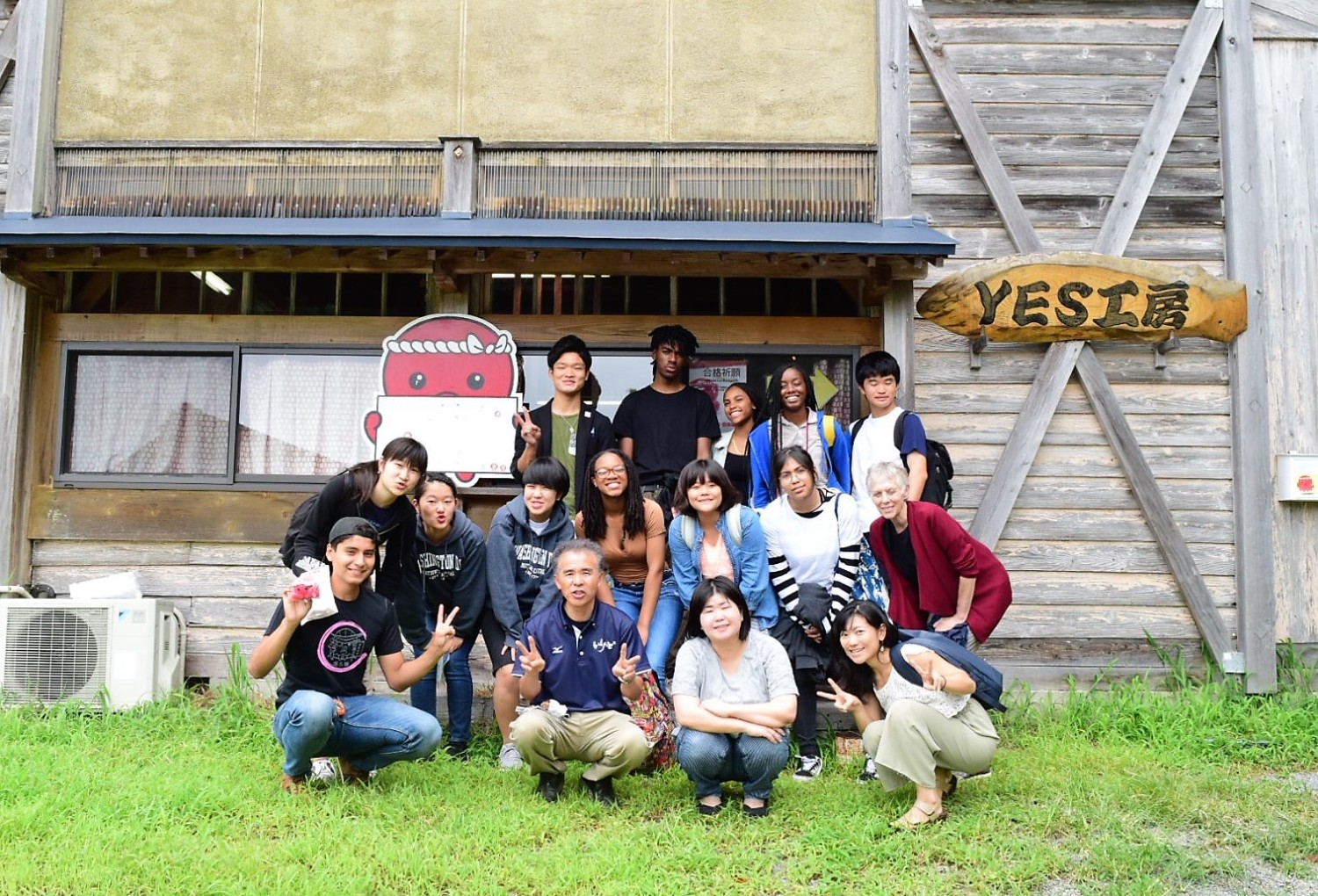 「YES工房」という木の看板がかかっている木造の建物の前で、国際色豊かな若者と大人が15人ほど並んでいる集合写真