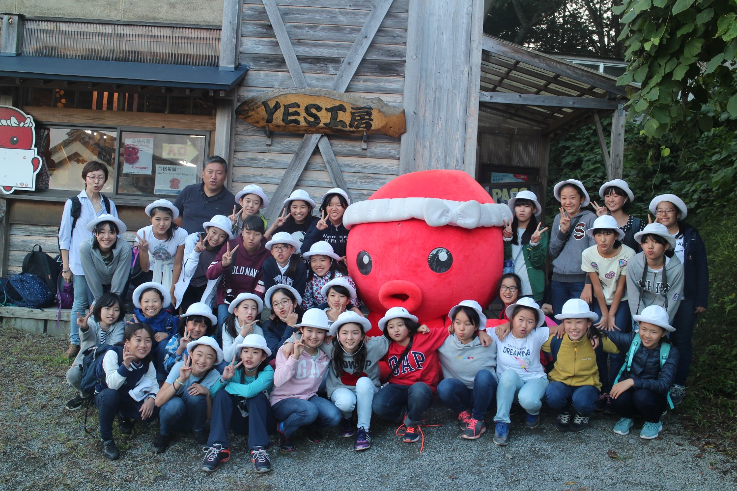 「YES工房」と書かれた看板が設置された建物の前で、白い帽子をかぶった多くの子どもたちが大きな赤いオクトパス君のマスコットと一緒に集合写真を撮っている写真