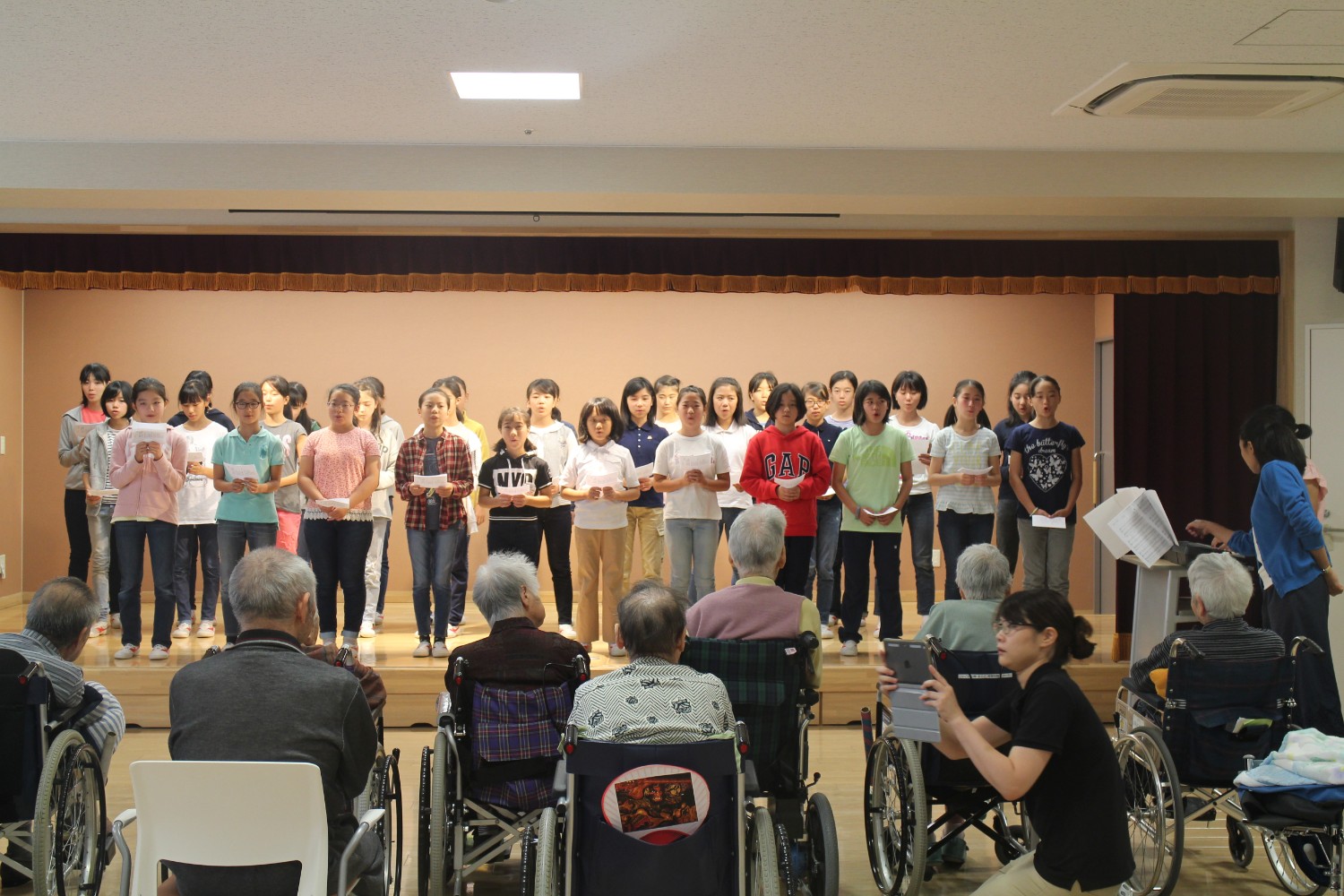 小さなステージに2列に並んだ児童の皆さんが歌詞カードを手にしながら高齢者の観客に向かって合唱を披露しており、椅子や車いすに座った高齢者たちが静かに耳を傾けている写真