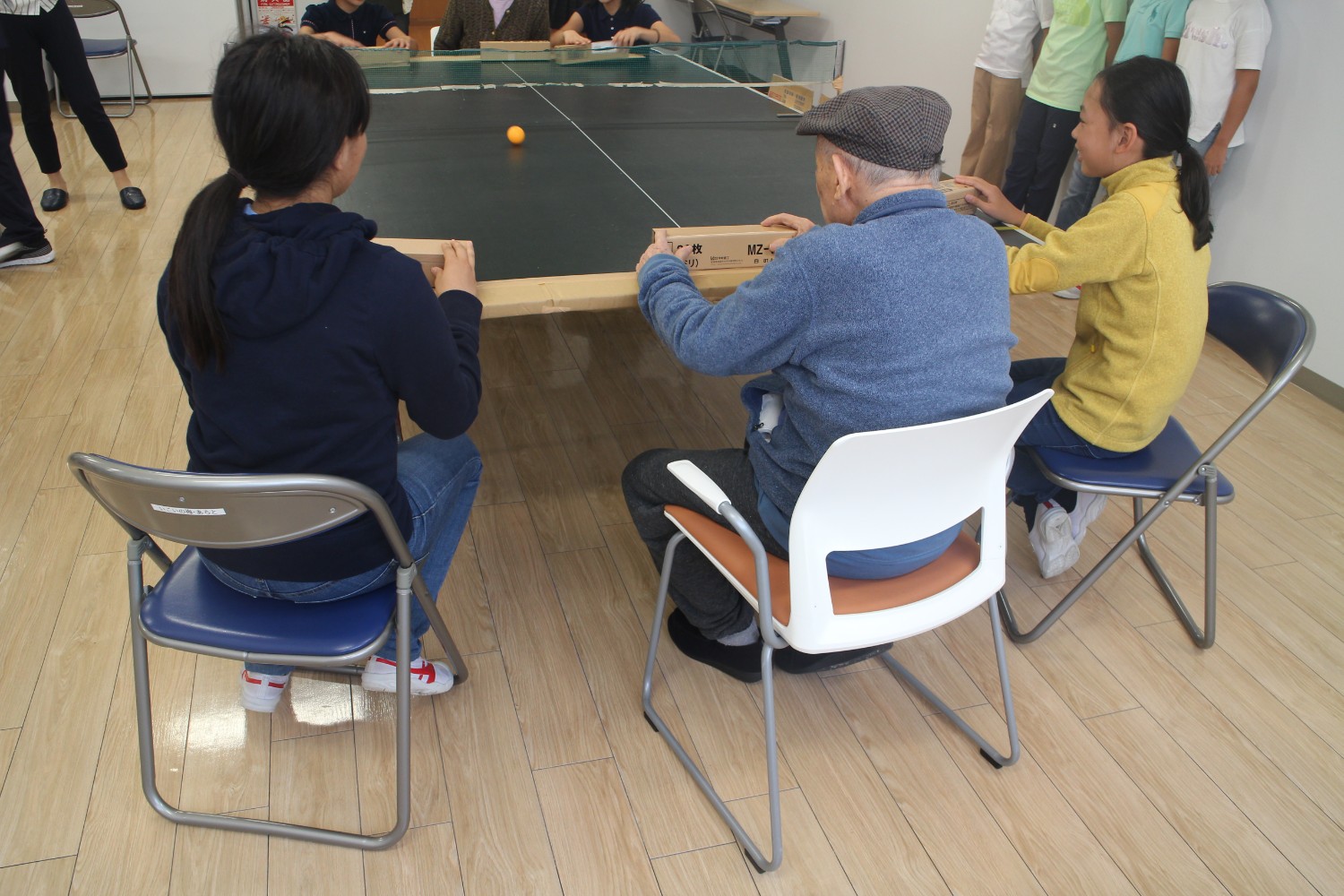 卓球台の端に椅子に座った二人の女子児童と高齢の男性が、段ボール製のパドルを使ってオレンジ色のピンポン球を打ち合っている写真