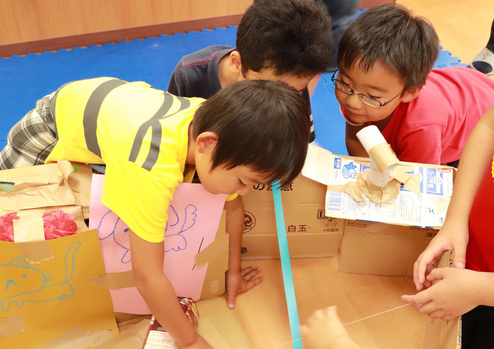 絵を描いた段ボールで3人の男の子が協力して家を作っている様子の写真