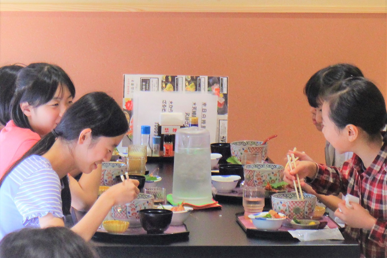 黒いテーブルに座った6人の女子児童たちが、和風の食事を囲んで笑顔で談笑しながら食事をしている写真