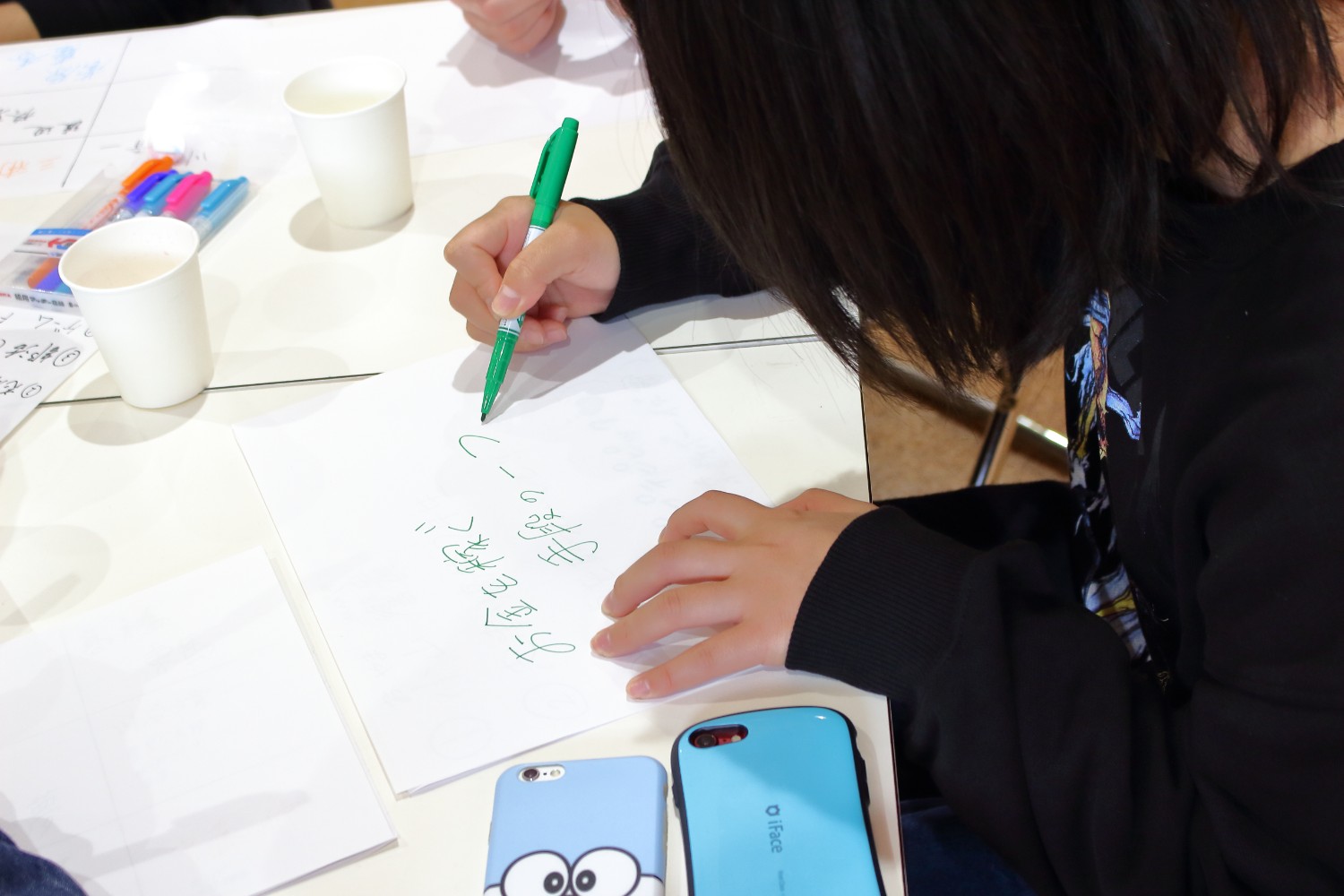 紙コップやスマートフォン、カラーペンが置かれた机の上の白い紙に「お金を稼ぐ手段の一つ」の文字を緑のペンで書いている黒いトレーナーを着た女性の手元を撮影した写真