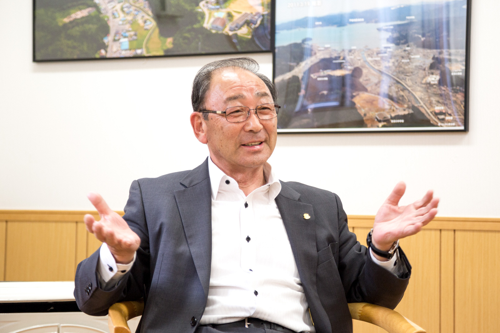 震災後の南三陸町の写真などが壁に飾られた部屋で、椅子に座り両手を広げてジェスチャーを交えて笑顔で話をしている遠藤さんの写真