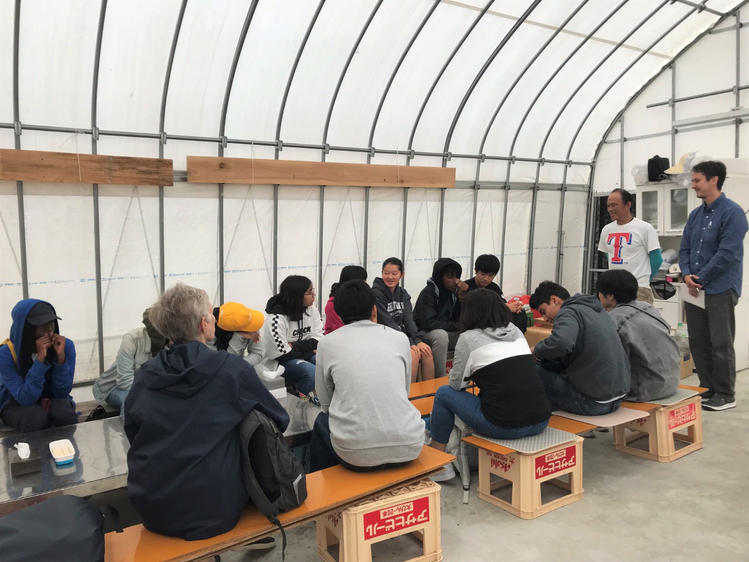 ビール瓶ケースと板で作られた簡易椅子が設置されたビニールハウスの中で、ベンチに座った十数人の若者たちが、前方に立っている男性の話を聞いている写真