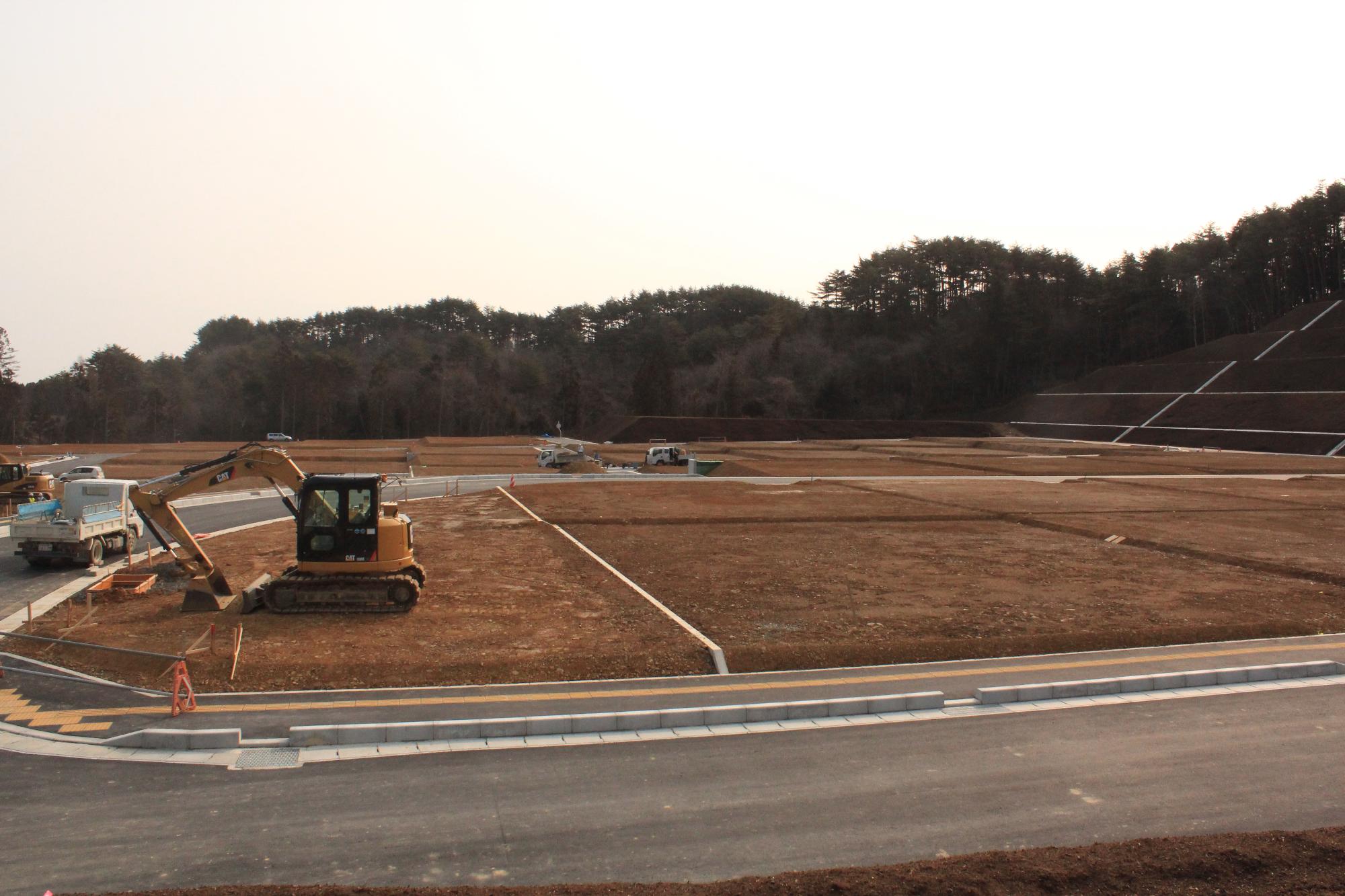 整地された造成地に重機と作業車が配置され、周囲の山林を背景に工事が進められている写真