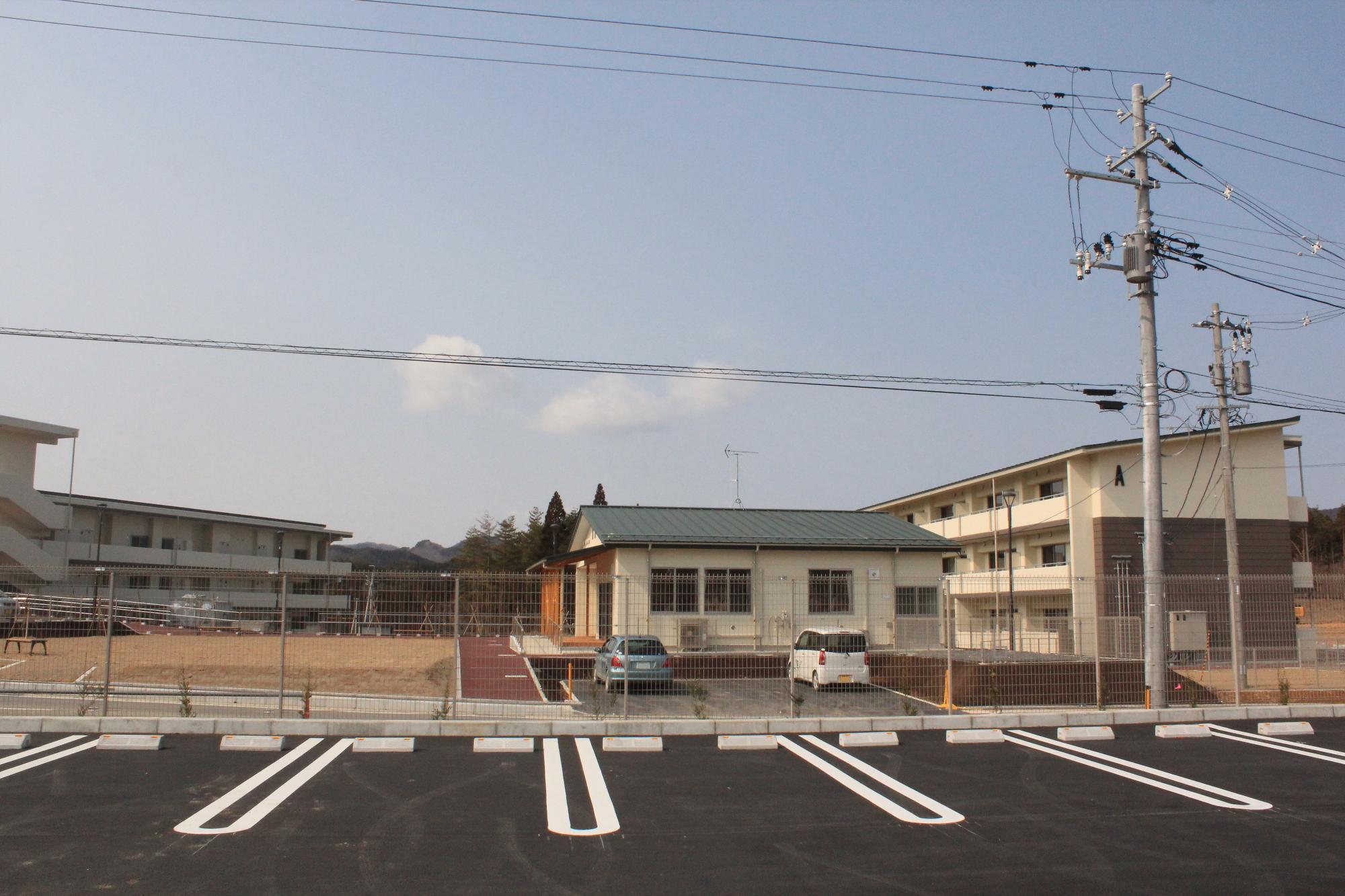 整備された新しいアスファルト駐車場の奥に、フェンスで囲まれた平屋の建物と中層集合住宅が並び、電柱や電線が空を横切っている様子の写真