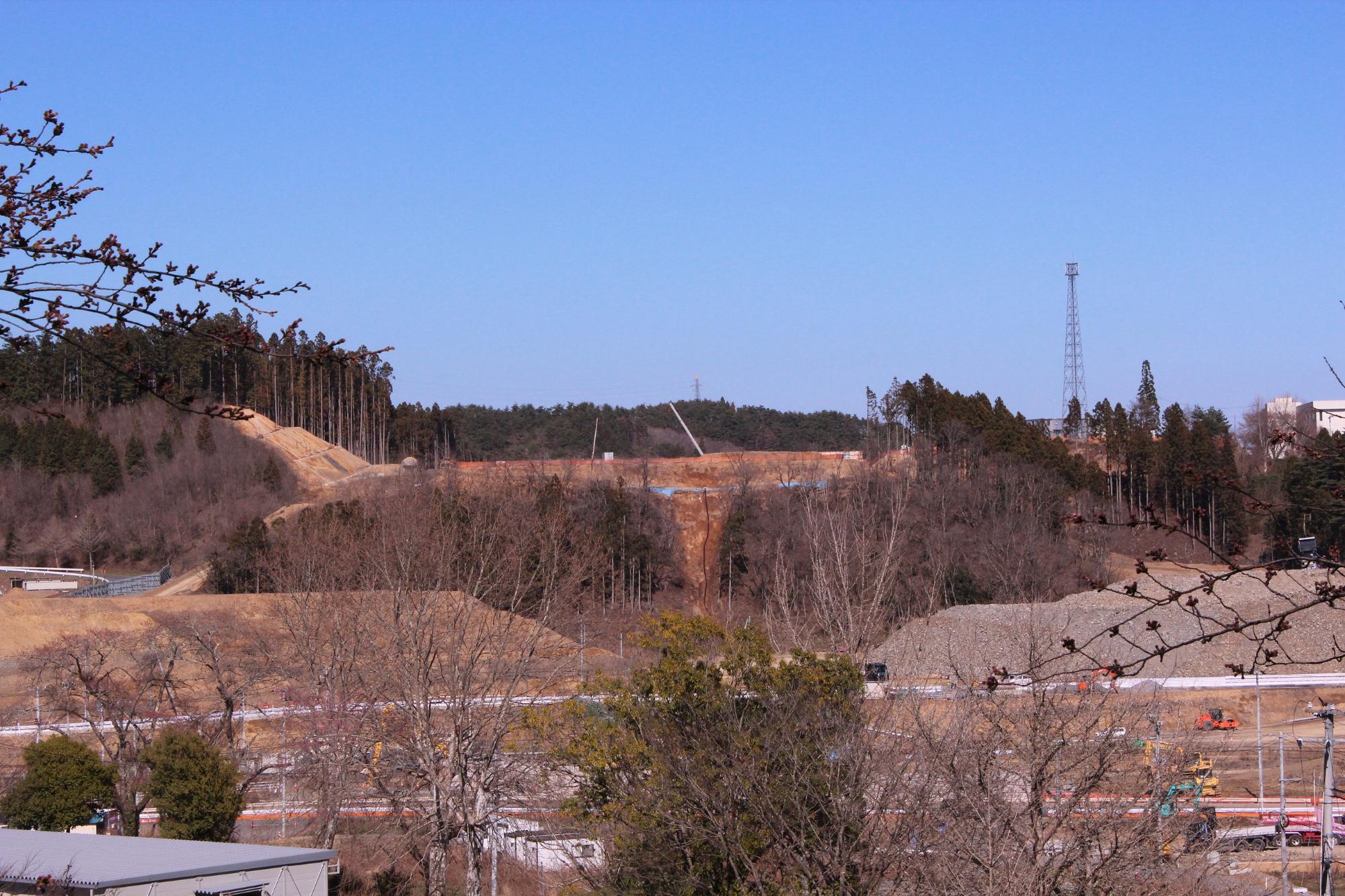 葉が落ちた枝の奥に造成地が見え、澄み切った青空の下造成地で2台のクレーン車が作業をしている様子がうかがえる写真