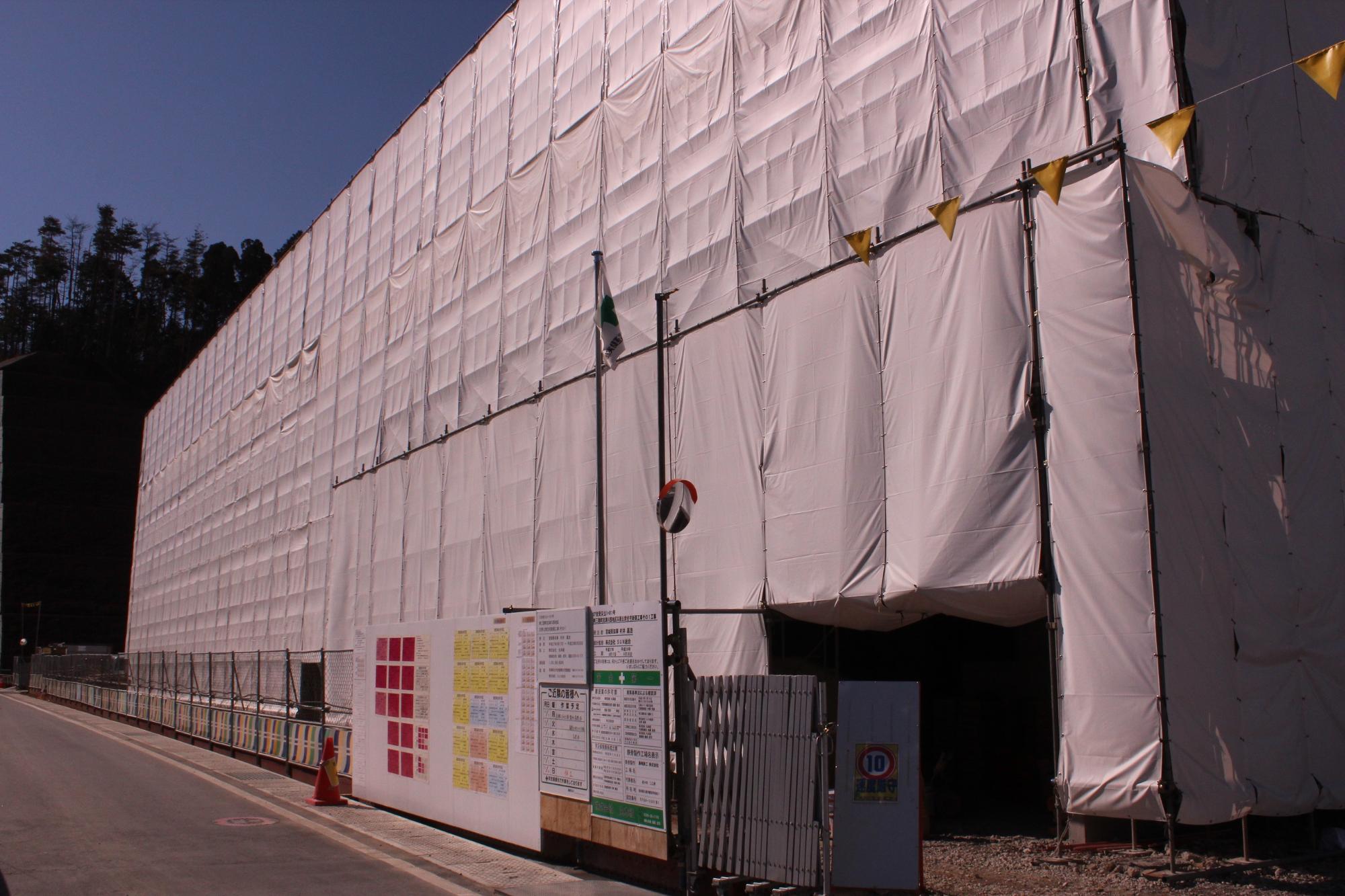 白いシートで覆われた大規模な建設現場の正面に、注意書きや作業予定表などが掲示された仮囲いが並び、入り口には安全旗や立て看板が設置されている写真