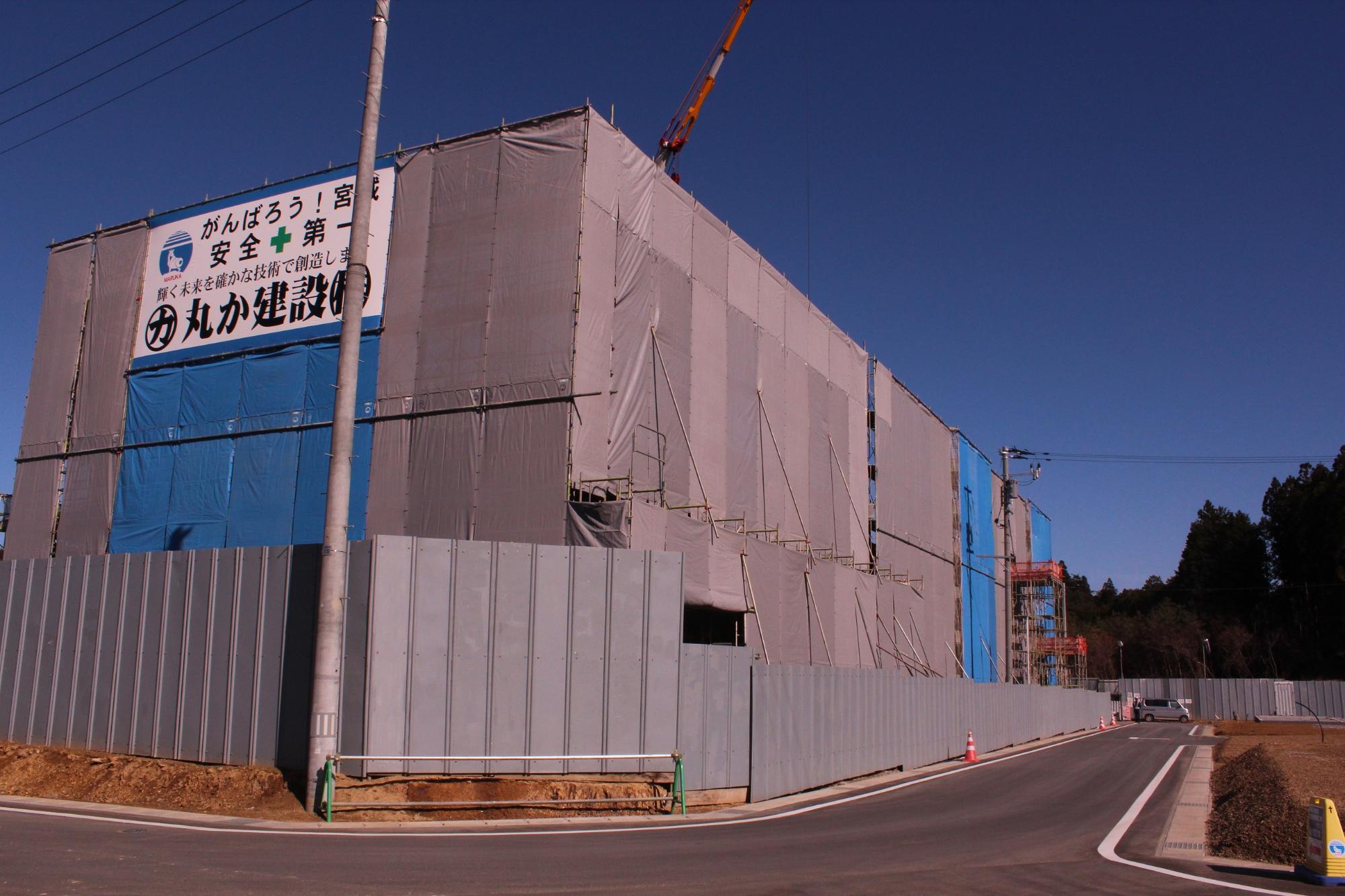 建設中の大きな建物がグレーと青のシートで覆われ、上部には「がんばろう！宮城 安全第一」と書かれた看板が掲げられ、建物周囲は高い仮囲いで囲まれている写真