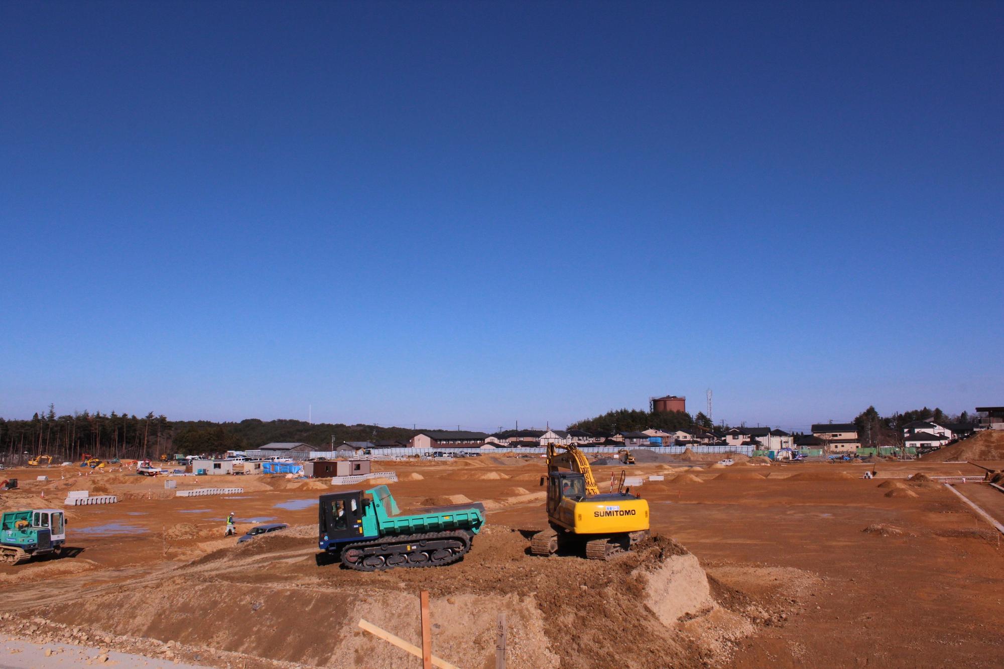広い青空の下、建設現場で複数の重機が茶色い土を掘削している様子が写っている写真