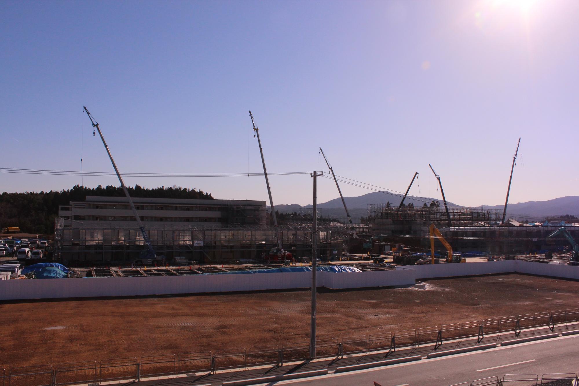 複数のクレーンが高く伸びる広大な建設現場で、左側に外観がほぼ完成した建物が立ち、右側では鉄骨と足場に囲まれた建物の建設が進み、多数の作業車両や資材が整然と配置されている写真