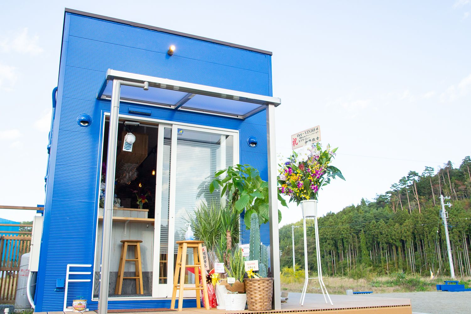 青いコンテナ型の建物の入り口に、開店祝いと思われる観葉植物や花が飾られており、ガラスの扉からは店内のカウンターと椅子が見えている写真