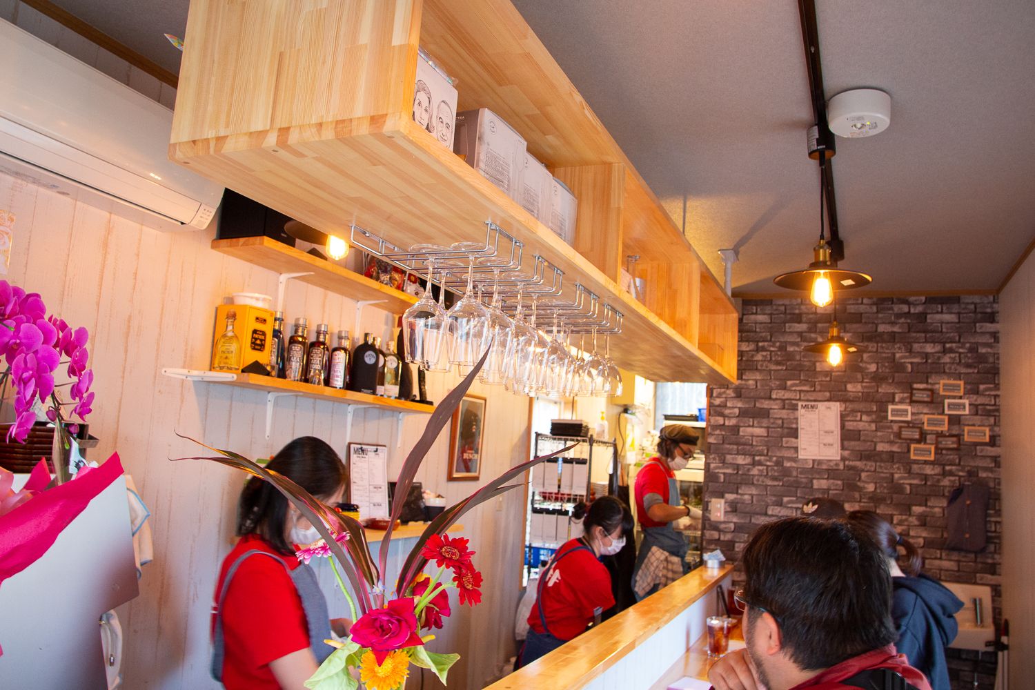 店内のカウンターに複数のスタッフが立ち、上部には吊り下げられたワイングラスや木製の棚がありカウンターにはお客さんが座っている写真