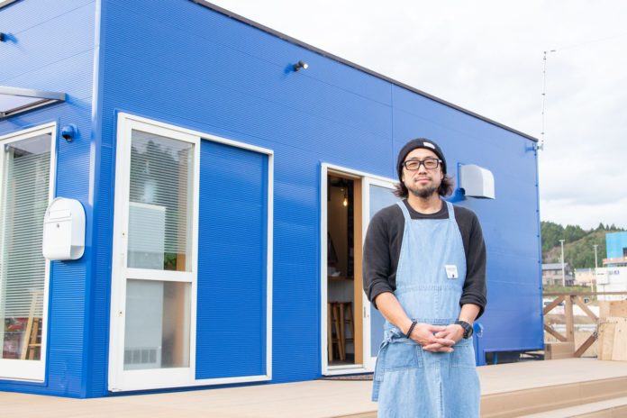 青いコンテナ型の建物の前で、黒いニット帽とメガネ、黒い長袖にデニムエプロンを着た男性が笑顔で立っている写真