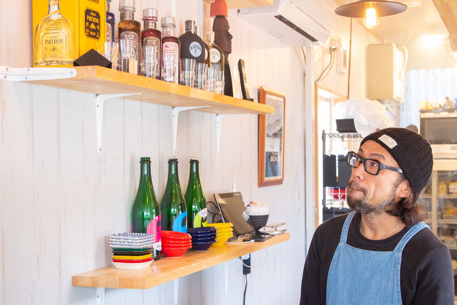 室内の白い壁に取り付けられた棚に、酒のボトルやグラス、小皿が並んでおり、その棚をエプロン姿の男性が見上げている写真