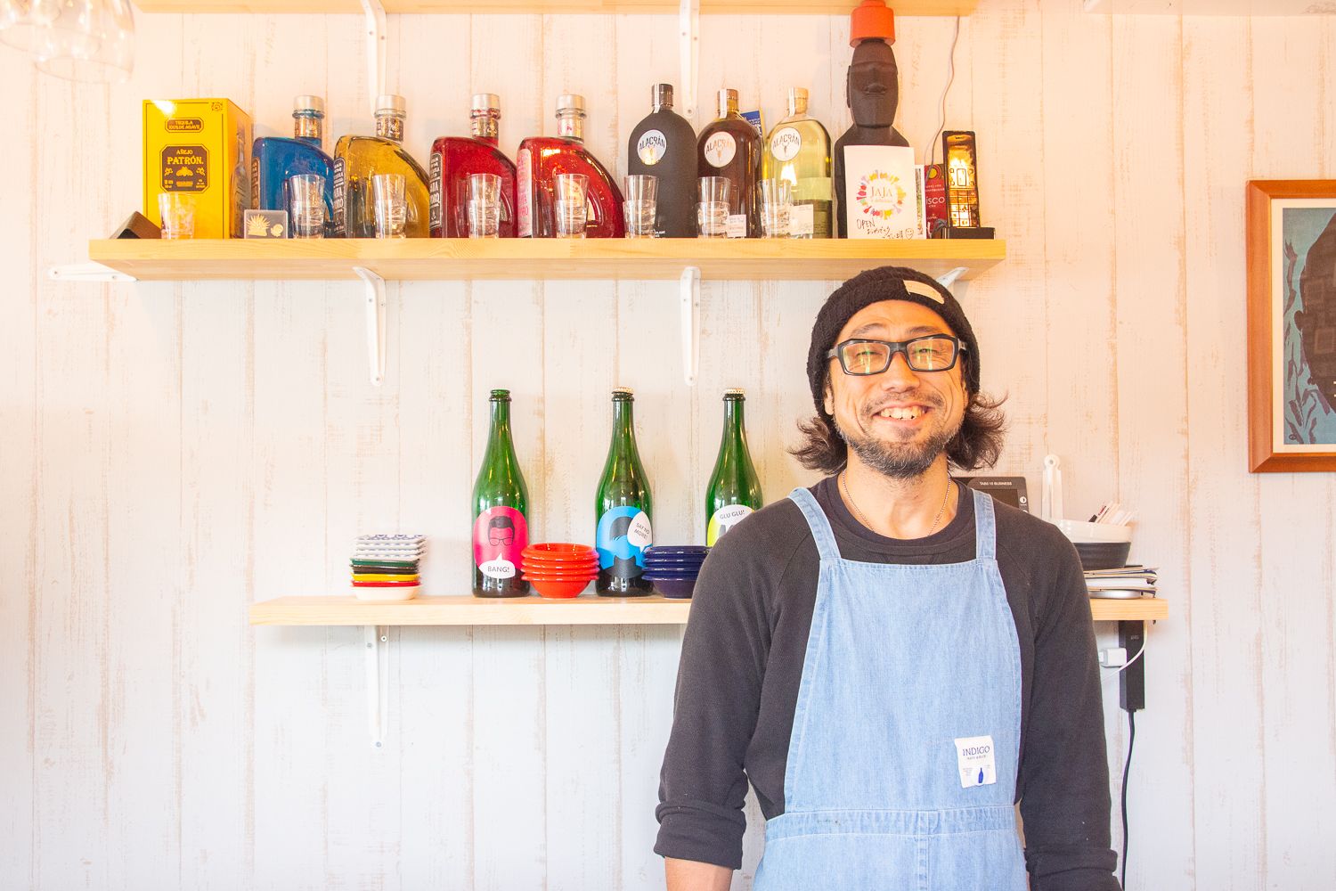 店内の白い壁の前で、デニムのエプロンを着た男性が満面の笑みを浮かべて立っており、背後の棚にはカラフルな酒瓶や小皿が整然と並べられている写真