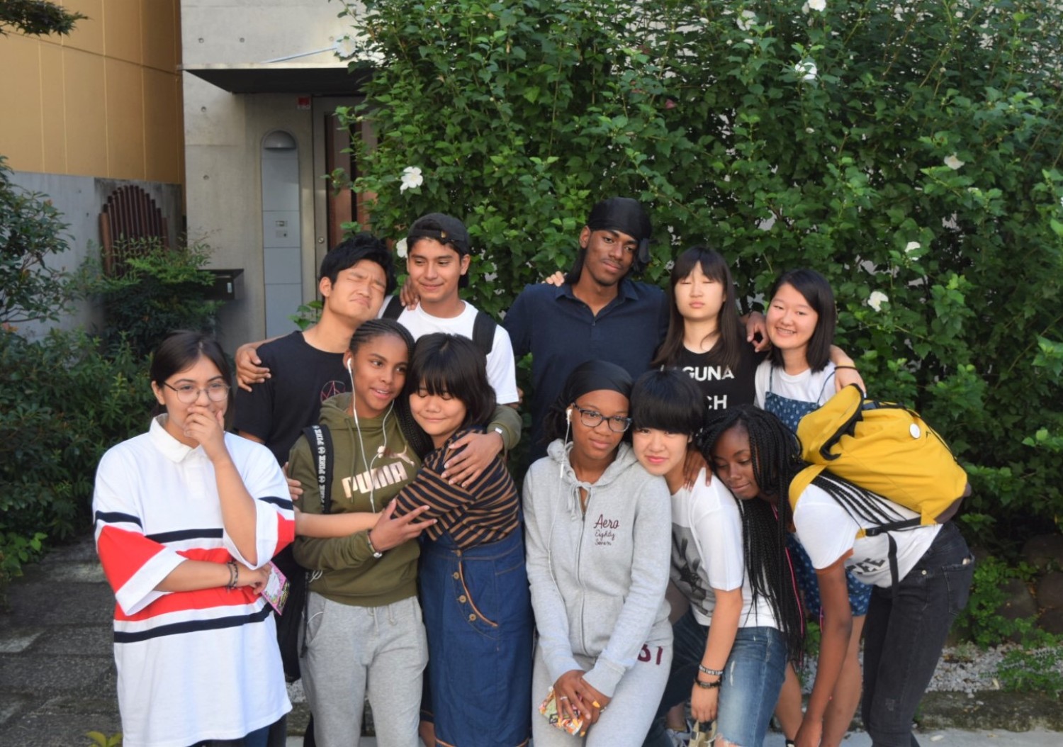 建物の前の屋外で木々を背景に、様々な服装の10代の若者たちが肩を寄せ合ってさみしそうな表情を浮かべ並んでいる写真