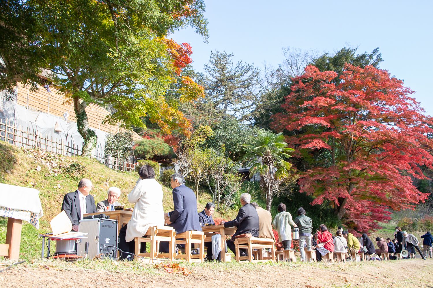 紅葉した木々に囲まれた自然の中で、長いテーブルと椅子を並べて大勢の人が屋外でお弁労を食べている写真