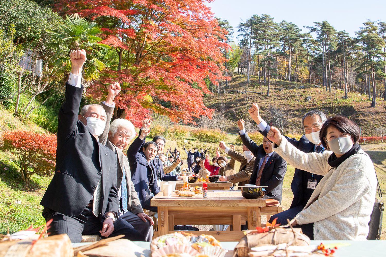 紅葉の山を背景に屋外で長テーブルを囲んだ人々が皆一斉に腕を高く上げてガッツポーズをしており、前には竹の皮に包まれた弁当が置かれている写真