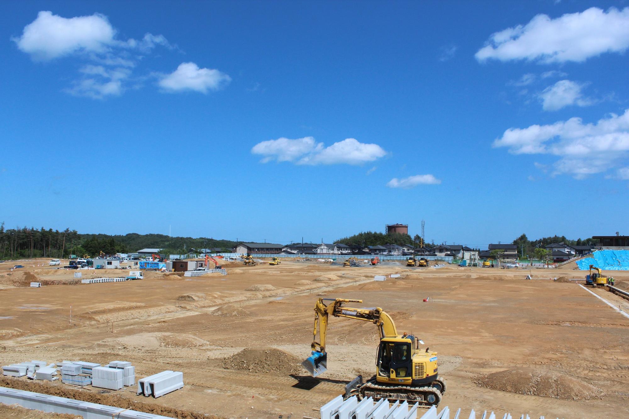 広い空き地で複数の建設機械が茶色い土の上で作業している様子が青空の下で撮影されている写真