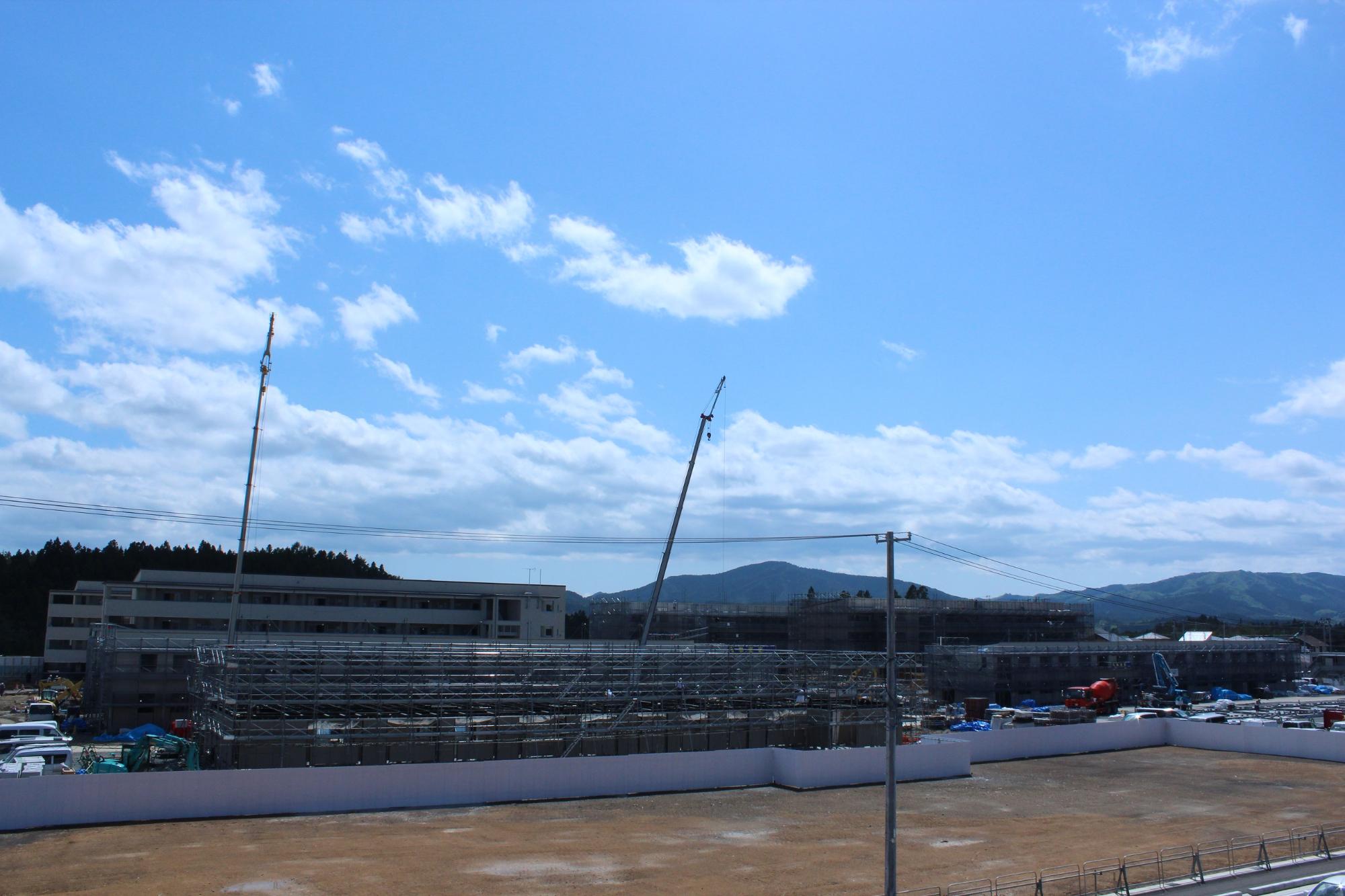 青空の下、左手に完成した四階建ての建物が並び、その手前と右奥では鉄骨の組み立てや外装工事が進行中で、複数のクレーンや建設機械が稼働している広大な建設現場の様子を捉えた写真