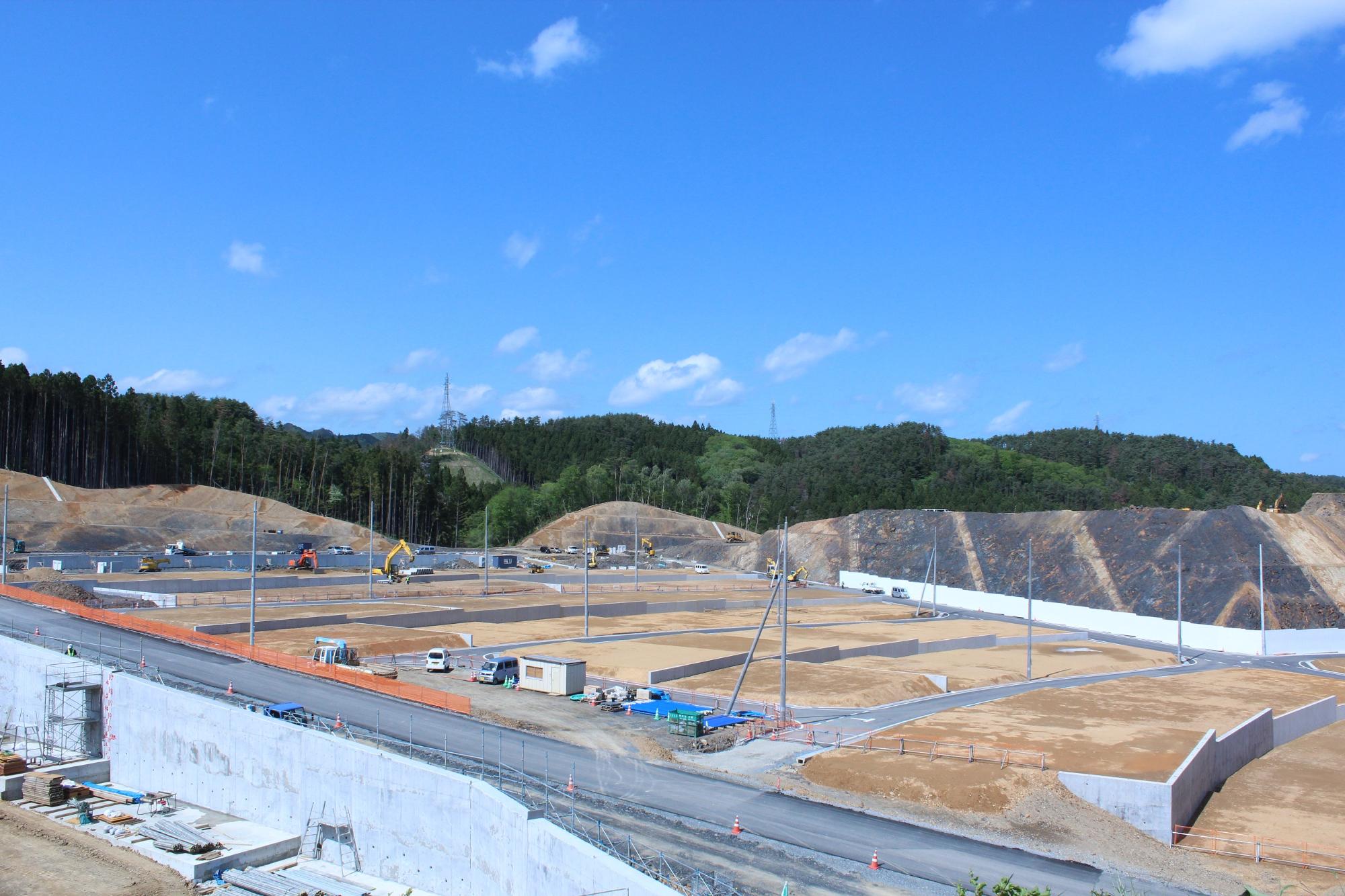 道路幅や曲線が明確に整いオレンジ色の仮設フェンスが設置され、ブルーシートでカバーされた資材置き場やロードローラーなどの重機が見え、各宅地区画の境界が整理される段階に入っている様子がうかがえる写真