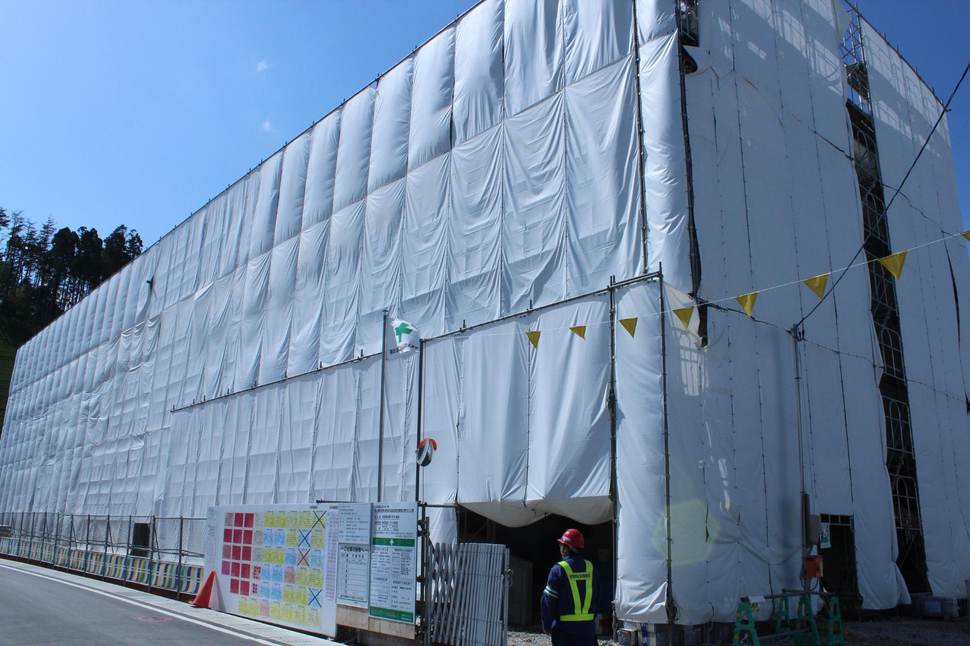白い防塵シートで全面が覆われた大きな建設中の建物の前に、ヘルメットと反射ベストを着た作業員が立ち、安全掲示板や黄色い安全旗が設置されている様子が写っている写真