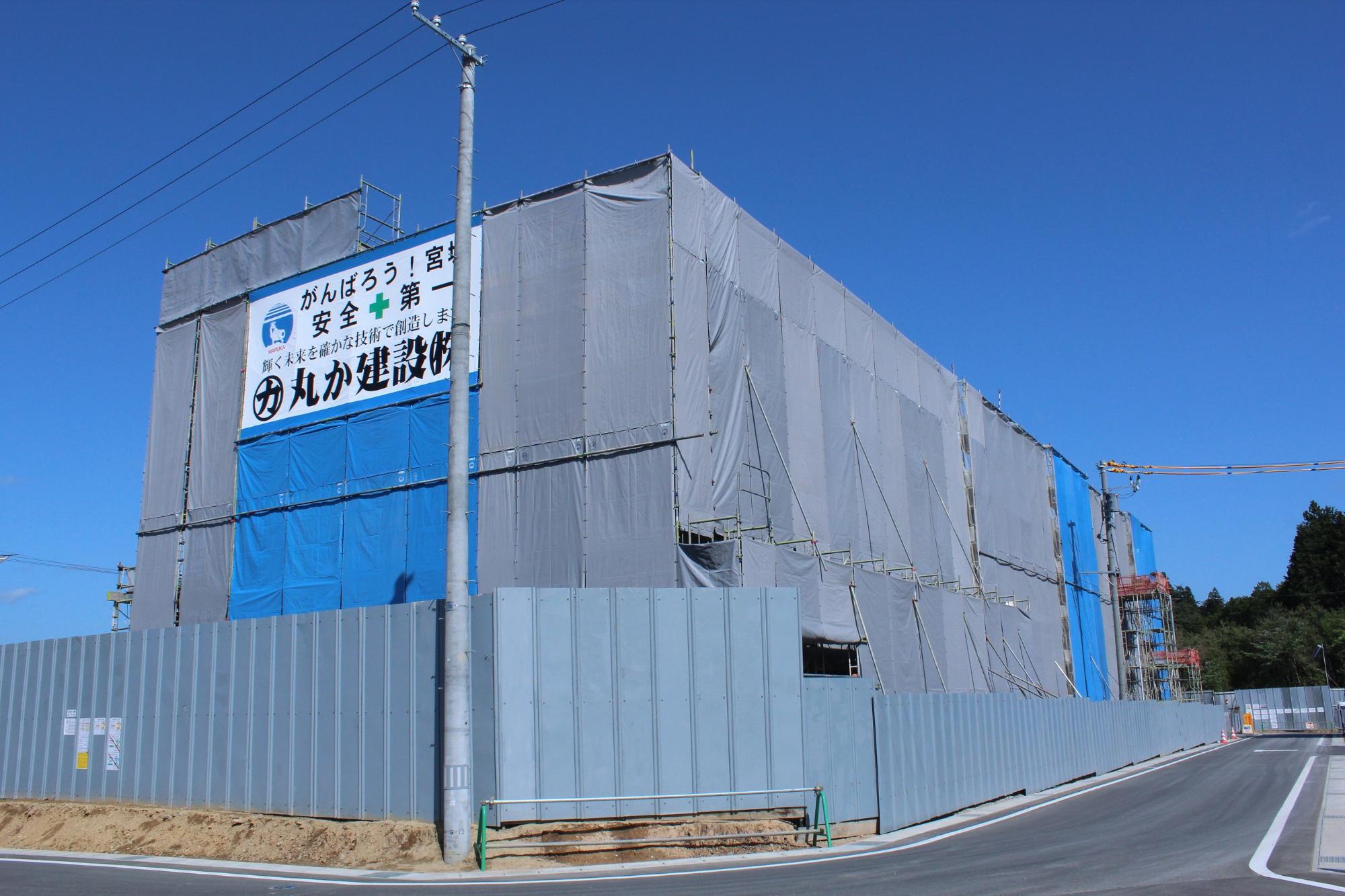 グレーと青の防塵シートで覆われた大規模な建設中の建物に、「がんばろう！宮城」や「安全第一」などの標語が大きく掲げられた看板が取り付けられており、その周囲が仮囲いで囲まれている写真