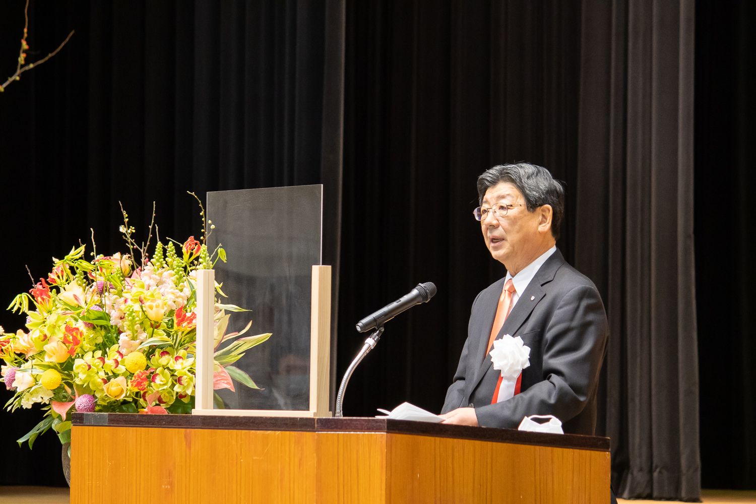 スーツを着てオレンジ色のネクタイをした佐藤町長が壇上でマイクの前に立ち、透明のアクリル板越しに成人式のあいさつをしている様子の写真