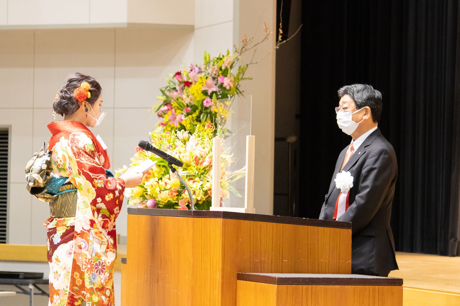 アクリル板が設置された檀上に立つスーツ姿の佐藤町長に向かって、赤と金を基調とした花柄の振袖を着た新成人代表の女性が、マイクの前で誓いの言葉を述べている様子の写真