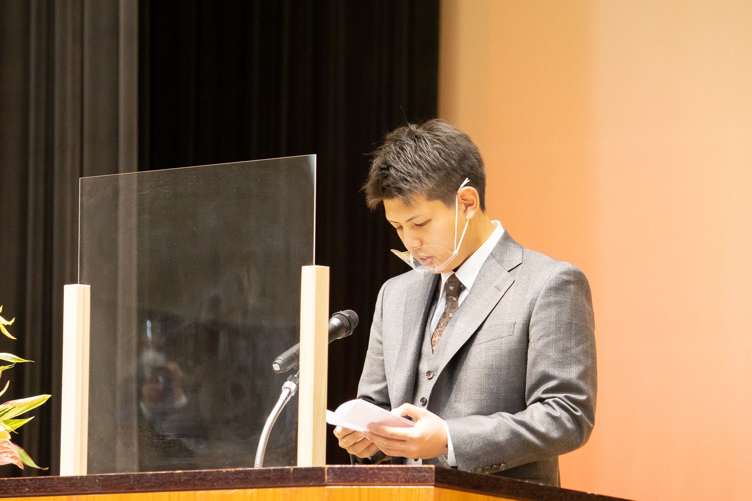 灰色のスーツを着た新成人の男性が、透明のアクリル板が設置された壇上でマイクの前に立ち、原稿を読みながら真剣な表情でスピーチしている様子の写真