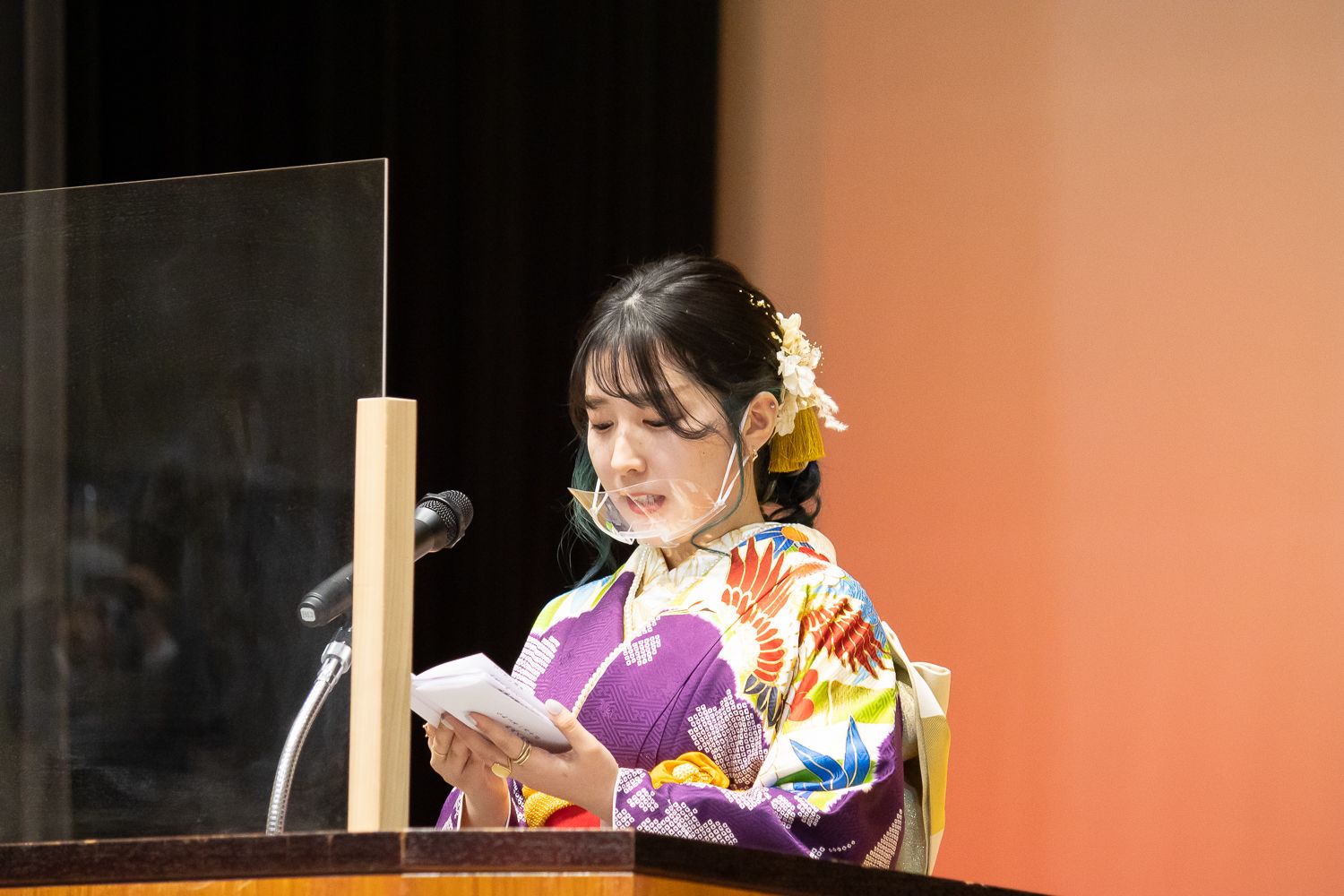 髪には白い花飾りがついており、紫色を基調とした華やかな振袖を着た新成人の女性が壇上で原稿を読みながらマイクの前でスピーチをしている写真