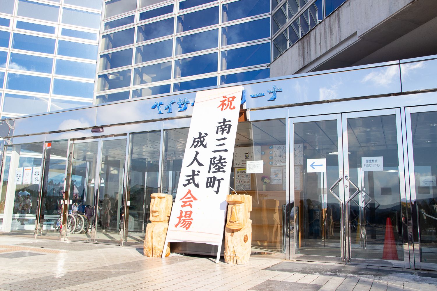 ガラス張りの近代的な建物の入口に木製のモアイ像が2体置かれ、間に「祝 南三陸町 成人式 会場」と大きく書かれた看板が立てられている成人式会場の入口の写真