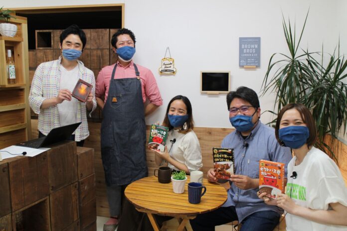 5人の男女がカフェで全員が青いマスクを着け座っている3人の方がカレーの箱を持ち、立っている1人の方が夕焼けの風景の写真を持って笑顔で写っている写真