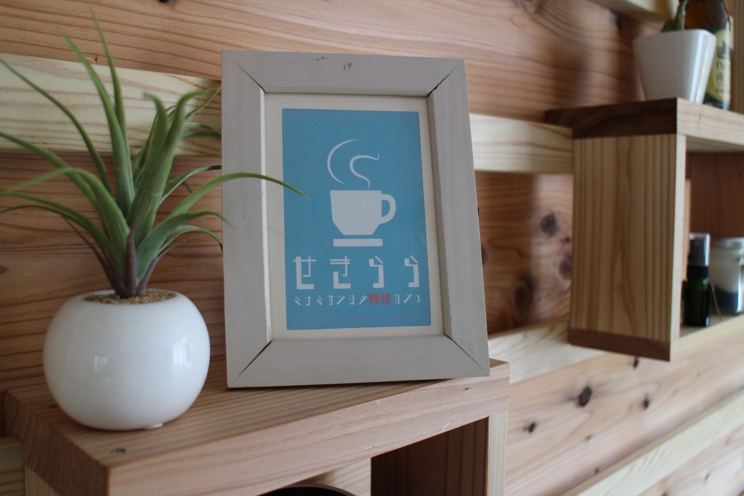 木の棚の上に小さな観葉植物と一緒に飾られている、水色の背景に湯気が立つコーヒーカップのイラストと「せきらら ミナミサンリク移住カフェ」の文字が書かれたポスターが白い写真立てに飾られた写真