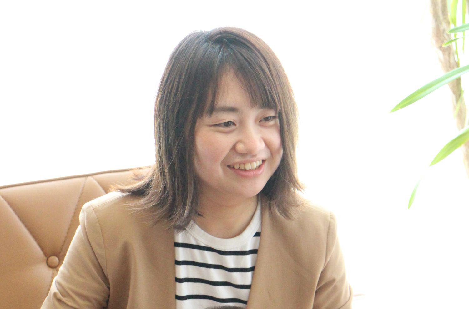 明るい室内で、ベージュのジャケットと白黒ボーダーのシャツを着た女性が椅子に座り、笑顔で誰かと話している様子を捉えた写真