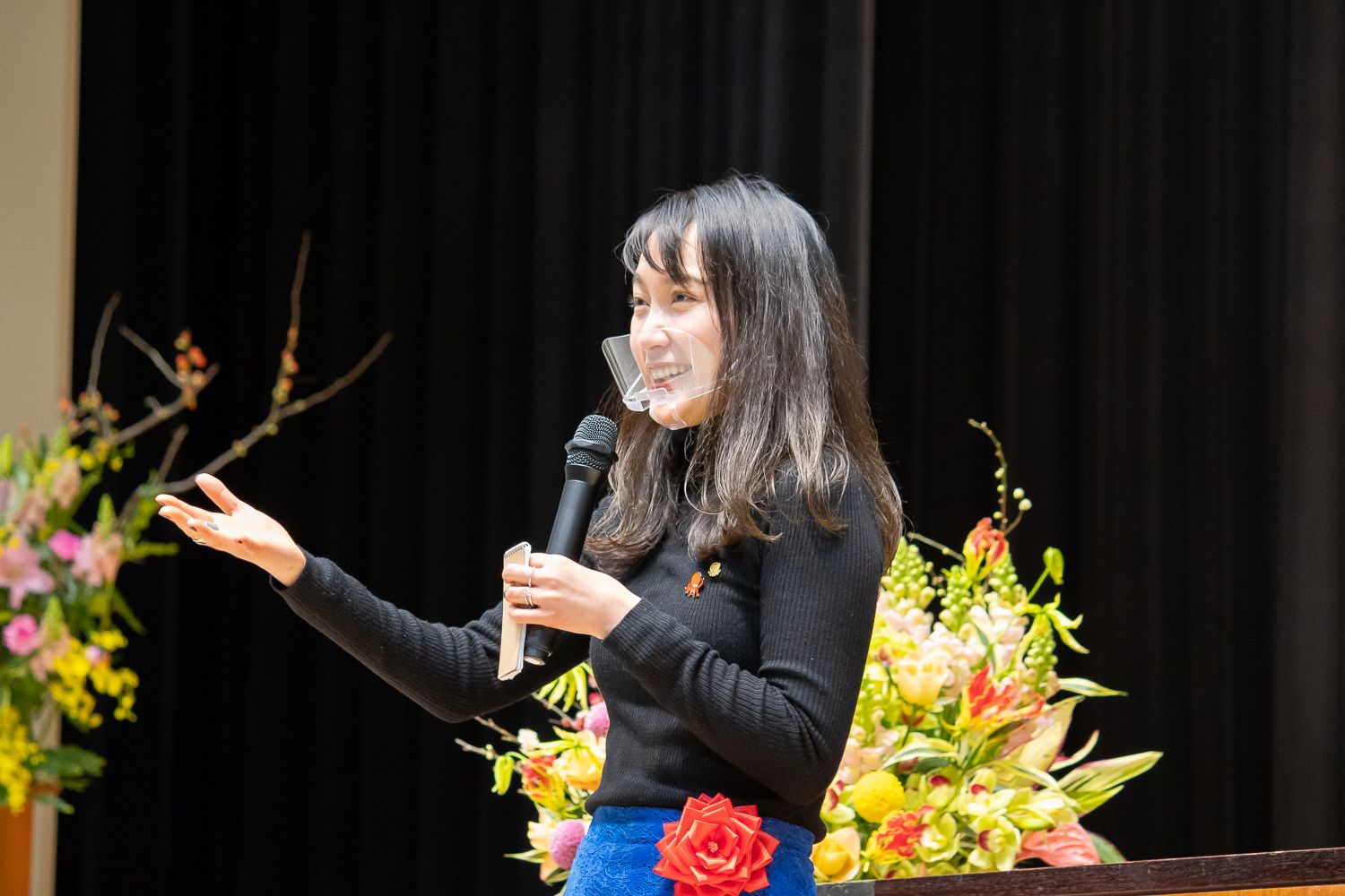 黒いニットと赤い花のコサージュをつけた青いスカートを着た女性が壇上でマイクを持ち、笑顔で手を広げながら話している写真
