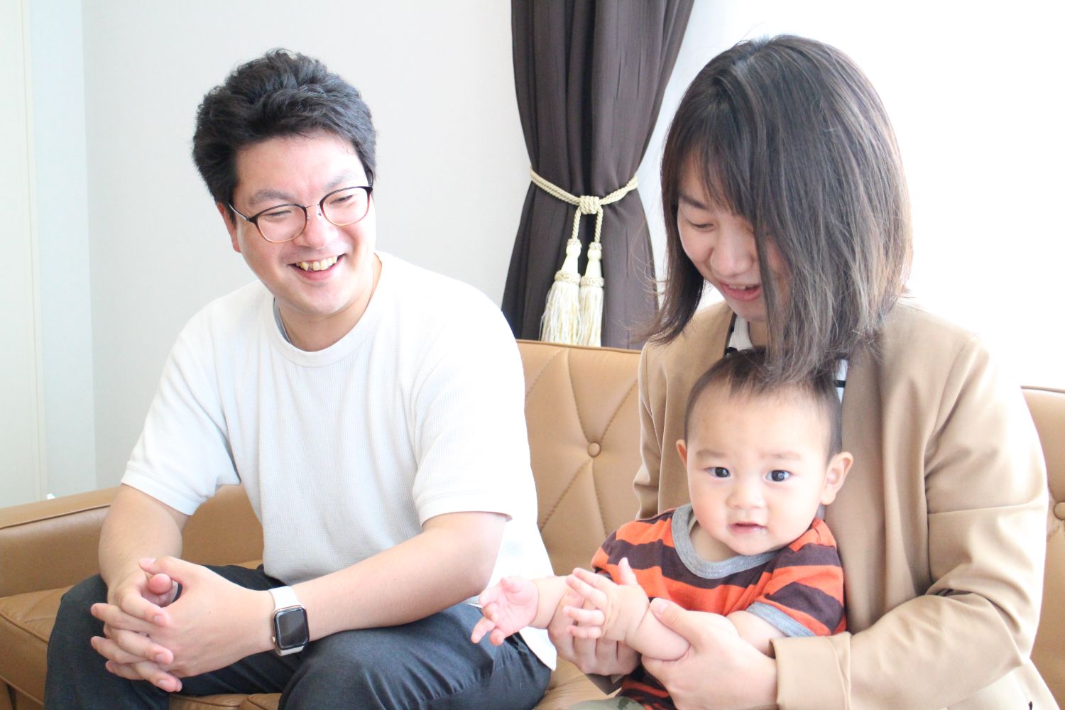 明るい室内で、白いシャツを着た佐藤さんが笑顔で見守る中、ベージュのジャケットを着た女性が膝に乗せた赤ちゃんを優しく抱きかかえ、赤ちゃんがカメラに向かってこちらを見つめている様子を写した写真