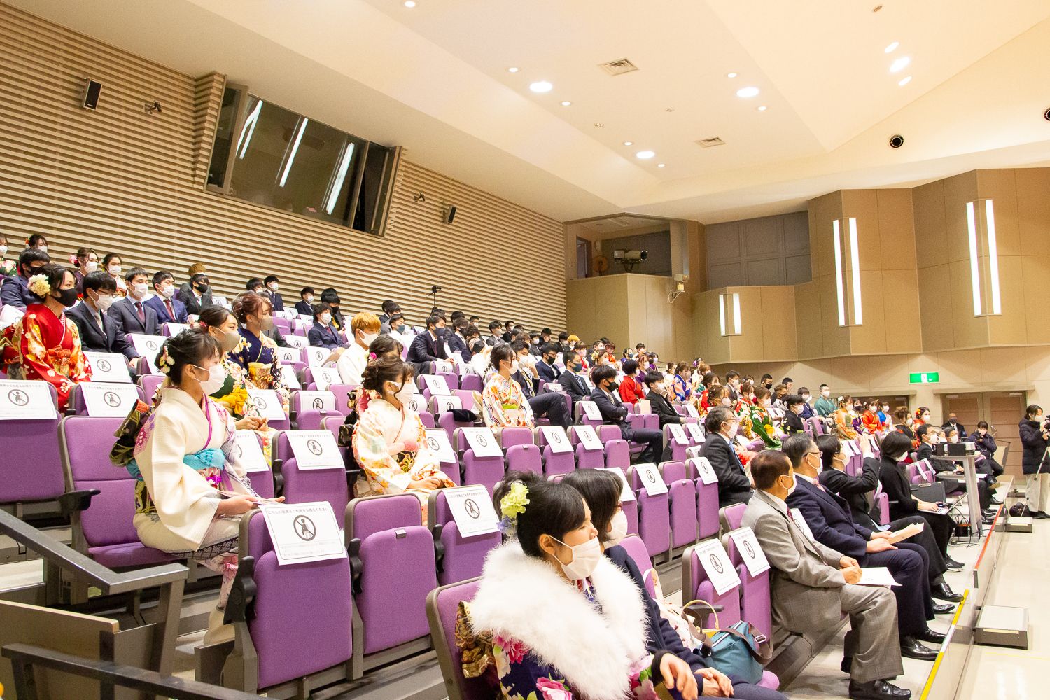 紫色の椅子が並ぶ会場で、新成人や関係者たちが間隔を空けて着席し、多くの女性は華やかな振袖姿で、男性はスーツ姿で式に参加している様子の写真