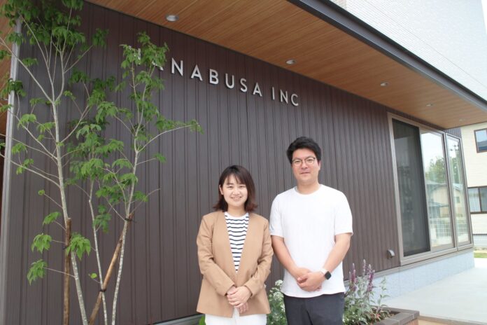 茶色い木目調の外壁に「HANABUSA INC」という看板が掲げられた建物の前で、ベージュのジャケットを着た女性と白いTシャツを着た男性が並んで立ち、カメラに向かって微笑んでいる様子を写した写真