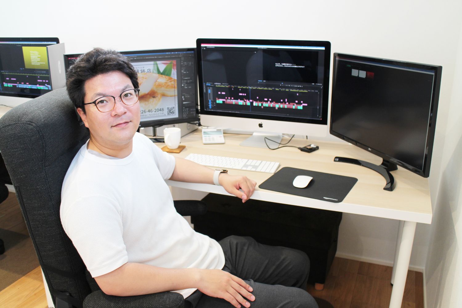 白いTシャツを着た男性が、映像編集ソフトが表示された複数のモニターに囲まれたデスクに座り、カメラに向かって穏やかに微笑んでいる様子を写した写真