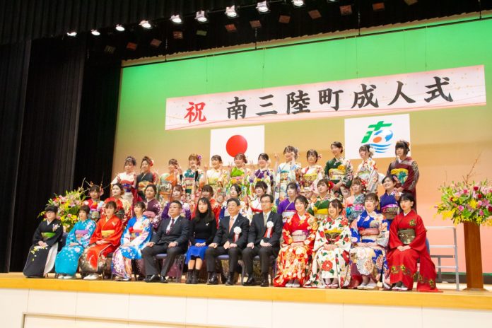 ステージ上で色とりどりの振袖を着た新成人の女性たちが3列に並び、中央に座ったスーツ姿の男性たちと一緒に記念撮影している成人式の集合写真