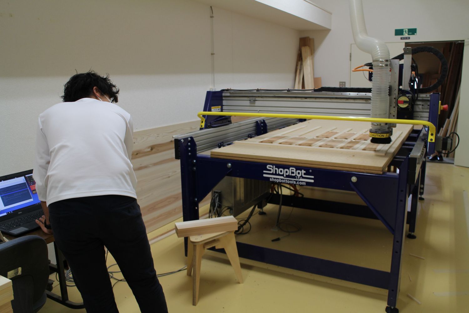 白い服を着た熊谷さんが木材が置かれた木材加工用の大型機械に向かってパソコンを操作しながら加工を行っている写真