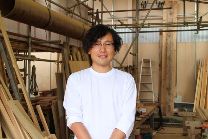 白い長袖シャツを着た熊谷さんが木材や工具が並ぶ木工所のような作業場で笑顔でカメラの前に立っている写真