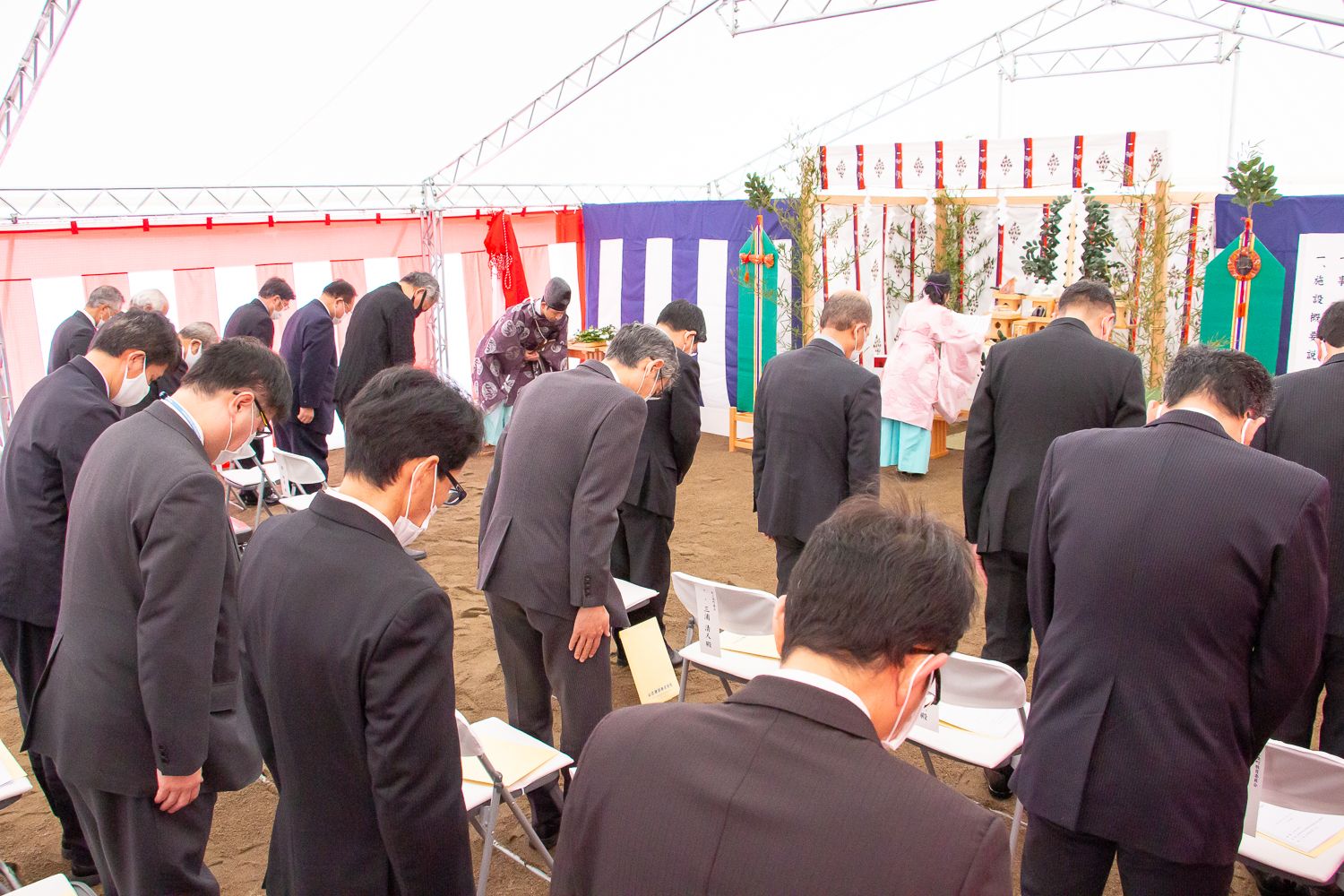 黒いスーツを着てマスクをしている複数の関係者が椅子から立ち上がり、神職の衣装を着た人物の前でお辞儀をしている式典の様子の写真