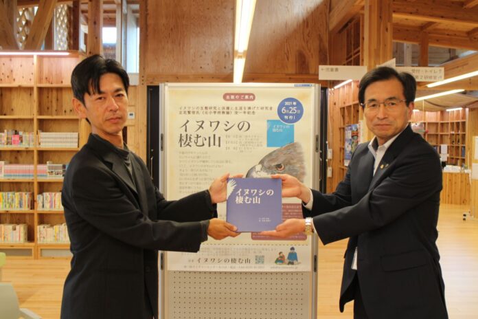 南三陸町生涯学習センターでスーツ姿の鈴木卓也さんと齋藤明さんが向かい合って1冊の本『イヌワシの棲む山』を共同で持ち、カメラに向かって紹介している様子を写した写真